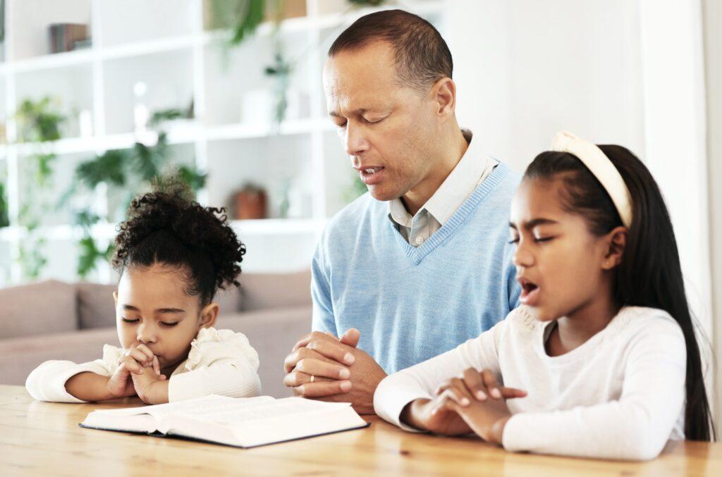 Family, worship and bible with father and kids praying at table for peace, religion and faith in th